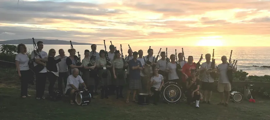 A crowd of pipers holding their pipes up at sunset by the ocean